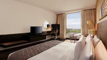 A hotel room with a large bed, a TV, a desk, a chair, a lamp, and a view of the city.