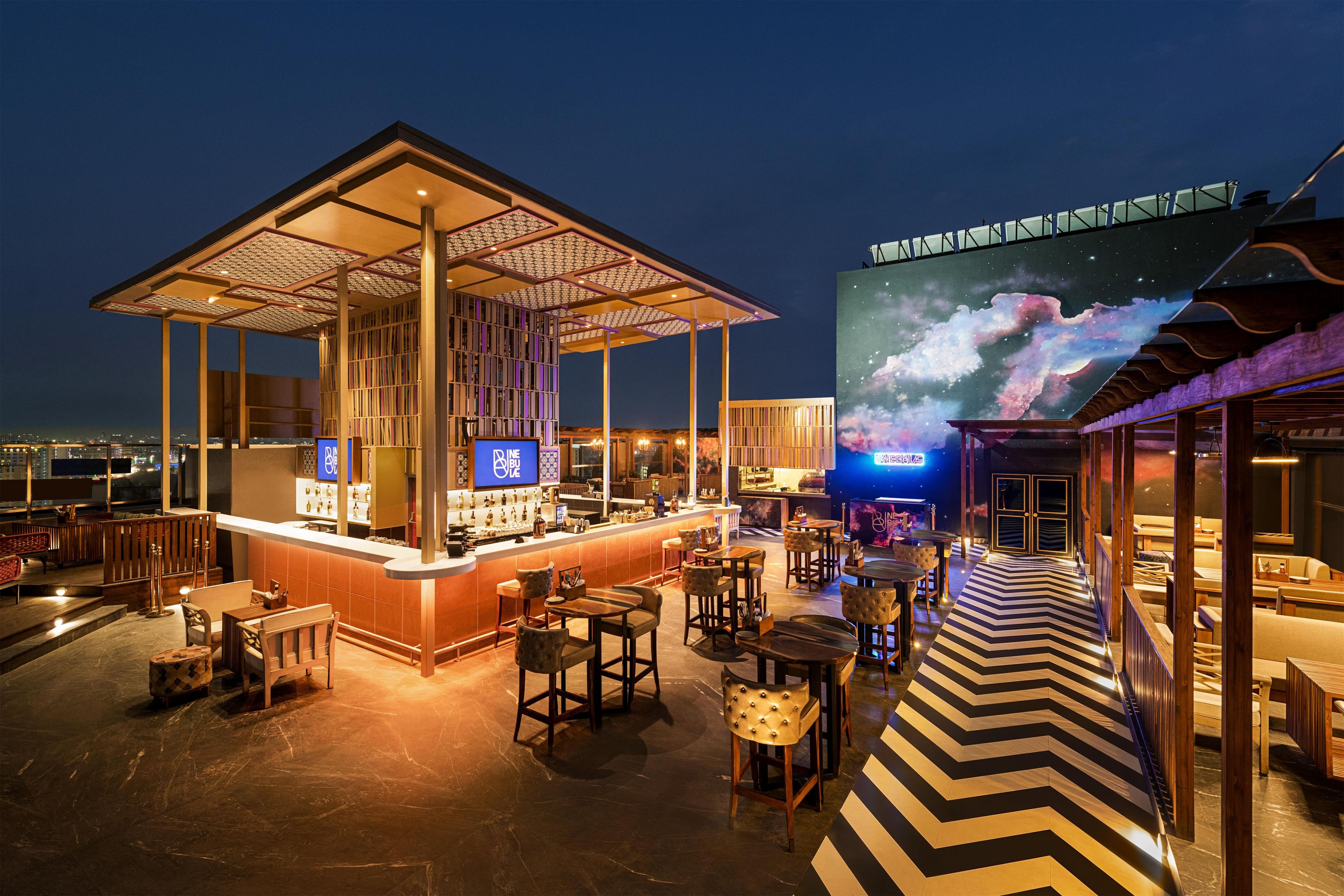 Un bar situé sur un toit, avec un comptoir, un espace salon et un grand écran numérique diffusant une scène cosmique.