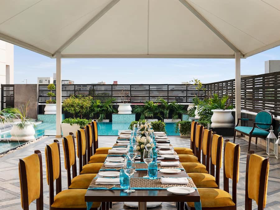 An outdoor dining area with a long table set for a meal, surrounded by chairs, with a pool and greenery in the background.