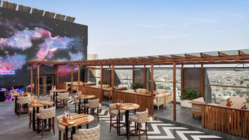 A rooftop bar with a city view, wooden furniture, and a large screen displaying a cosmic scene.