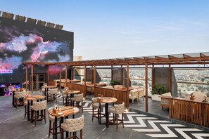 A rooftop bar with a city view, wooden furniture, and a large screen displaying a cosmic scene.