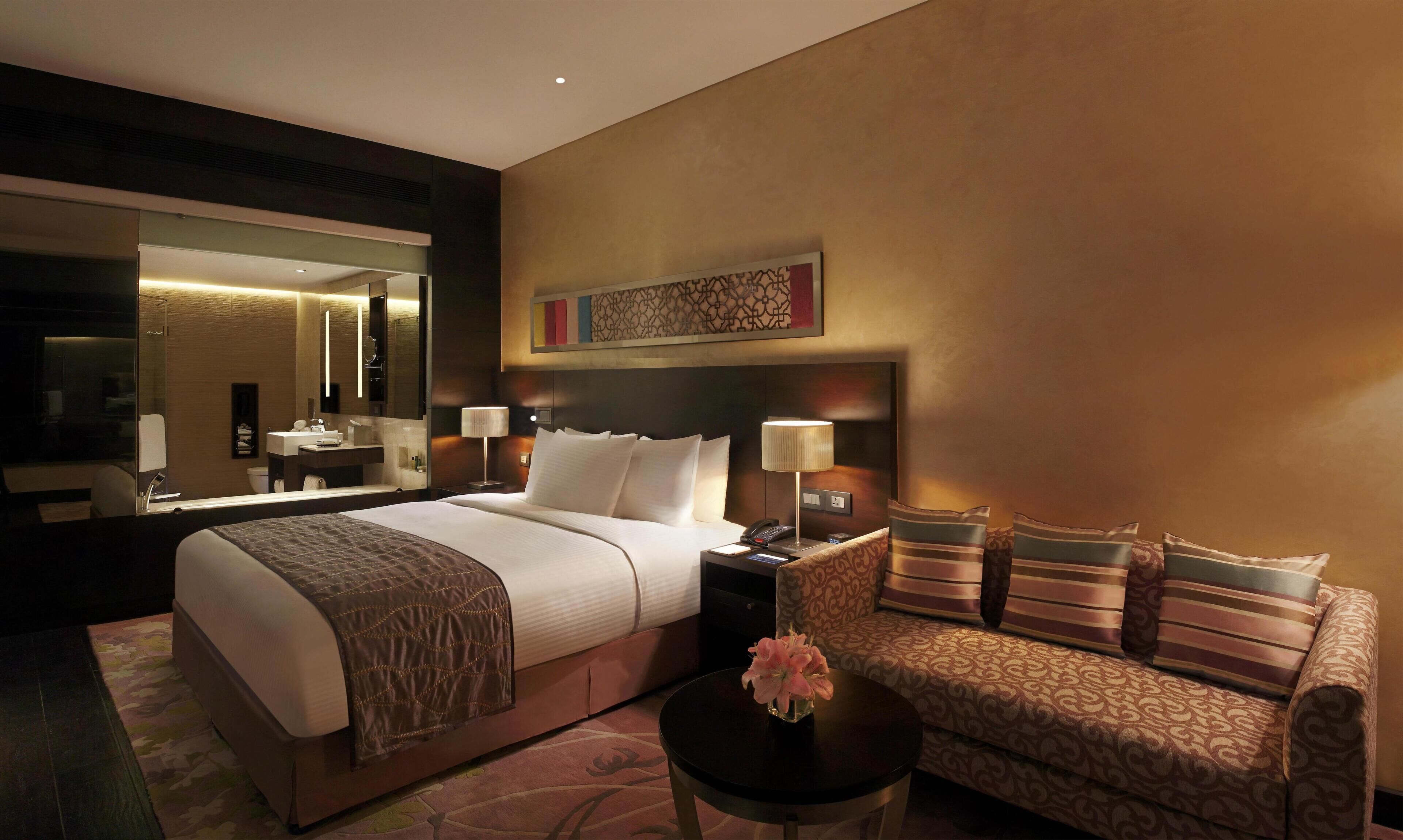 Une chambre d’hôtel avec un grand lit, un canapé, une petite table sur laquelle est posé un vase de fleurs, un miroir et un tableau au mur.