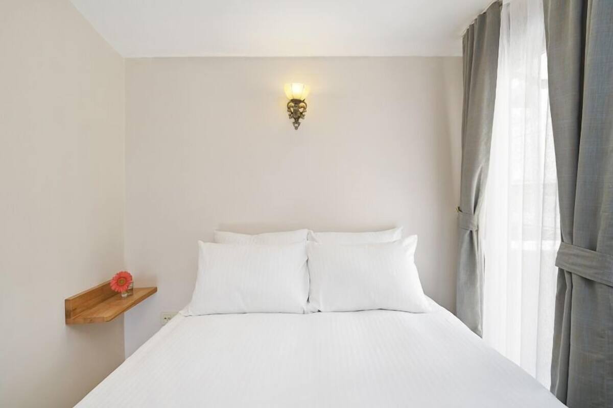 A neatly made bed with white linens, a wall-mounted light fixture, and a wooden shelf with a red flower.