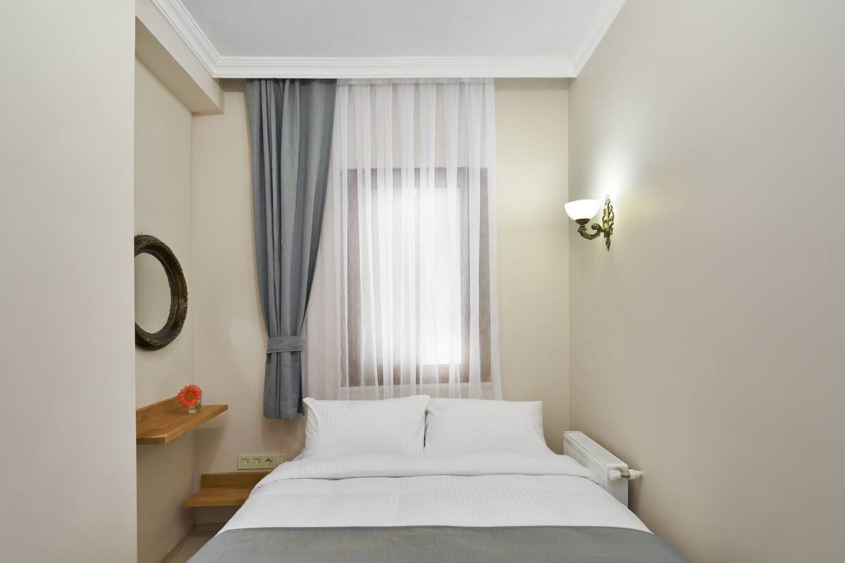 A neatly arranged bedroom with a bed, a bedside table, a mirror, and a window with sheer curtains.