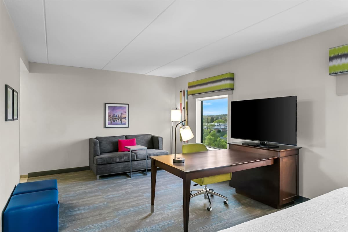 A hotel room with a desk, a television, a sofa, and a window with a view of greenery.