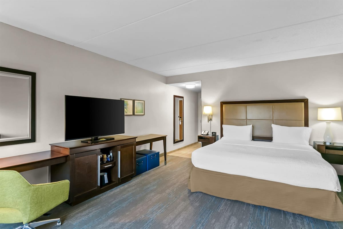 A hotel room with a large bed, a desk with a flat-screen TV, a chair, and a wardrobe.