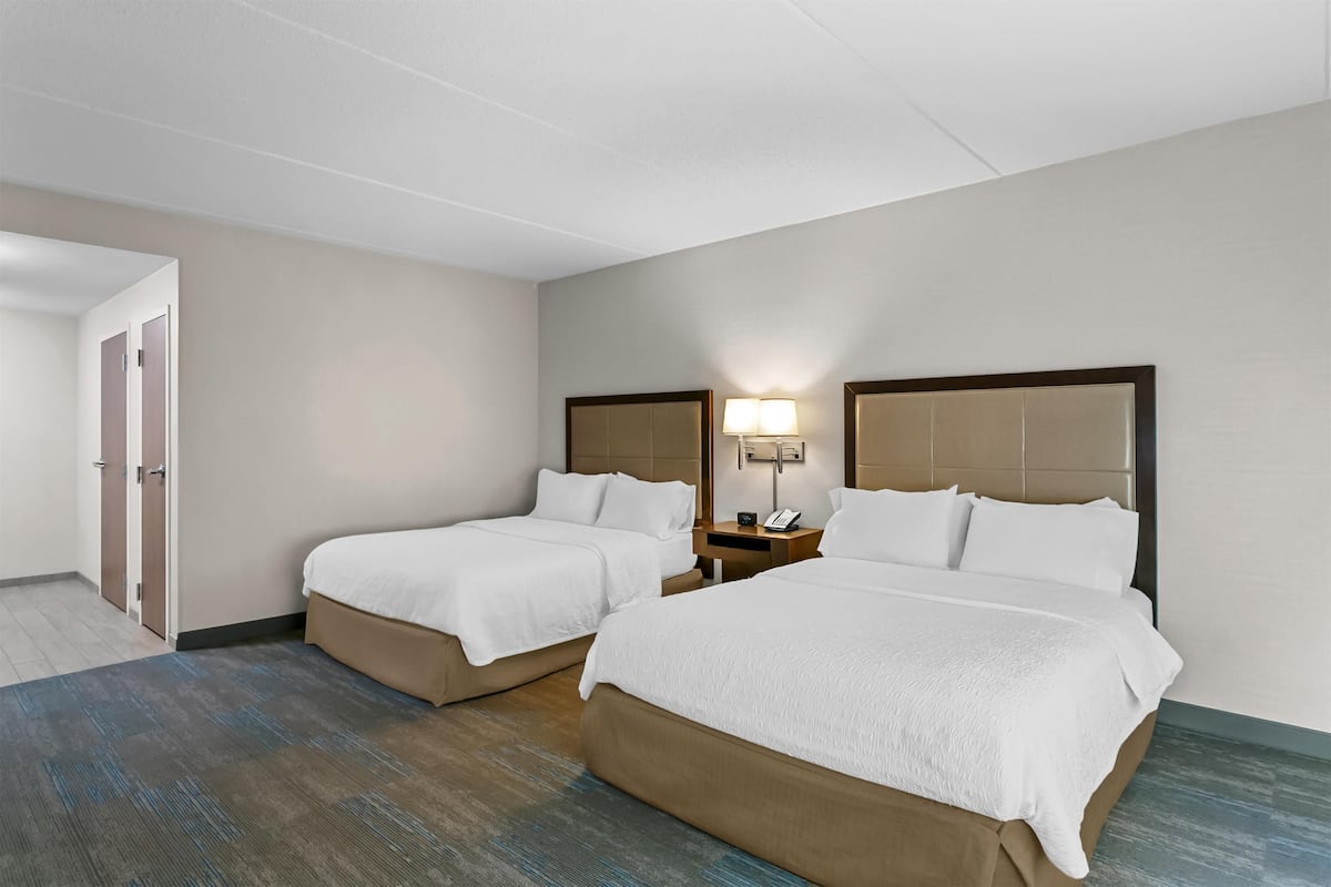 A hotel room with two beds, a bedside table with a lamp, and a door leading to another room.