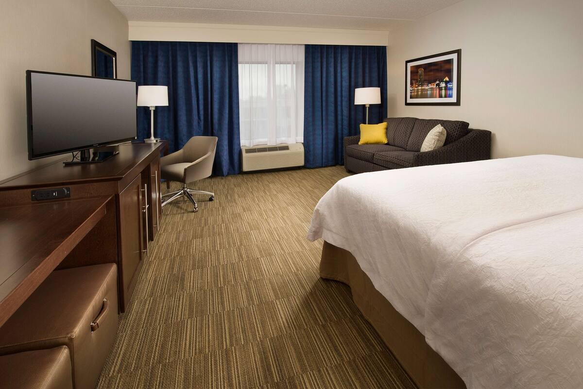 A hotel room with a bed, a desk with a TV, a chair, a sofa with a pillow, and a picture on the wall.