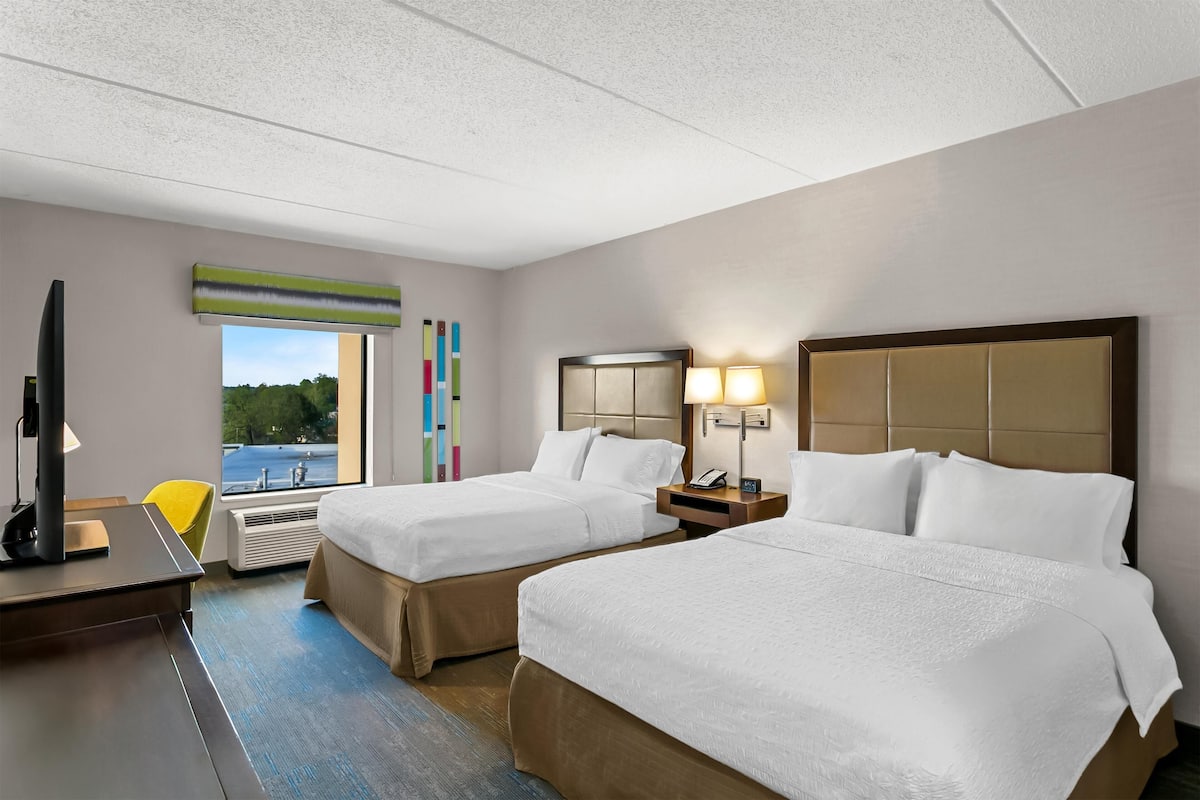 A hotel room with two beds, a desk, a chair, a TV, and a window with a view of trees.