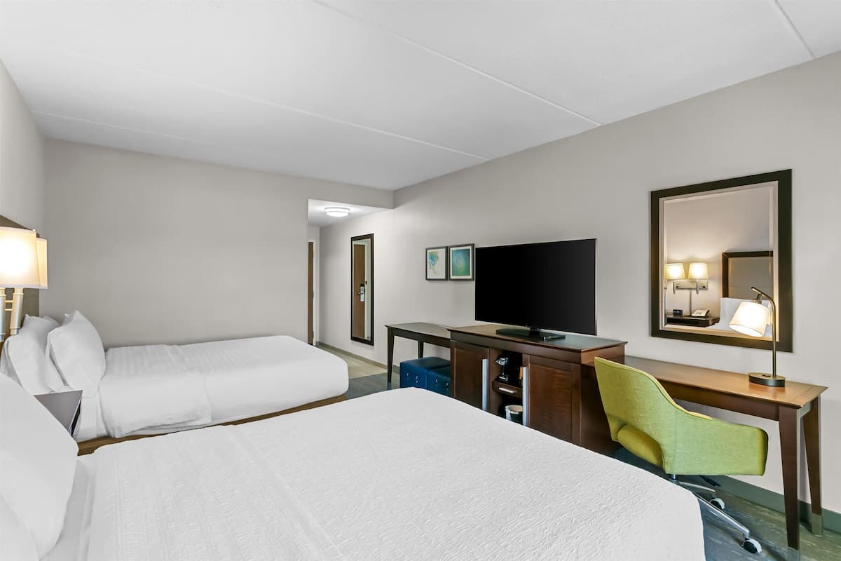 A hotel room with two beds, a desk with a television, a chair, and a mirror.