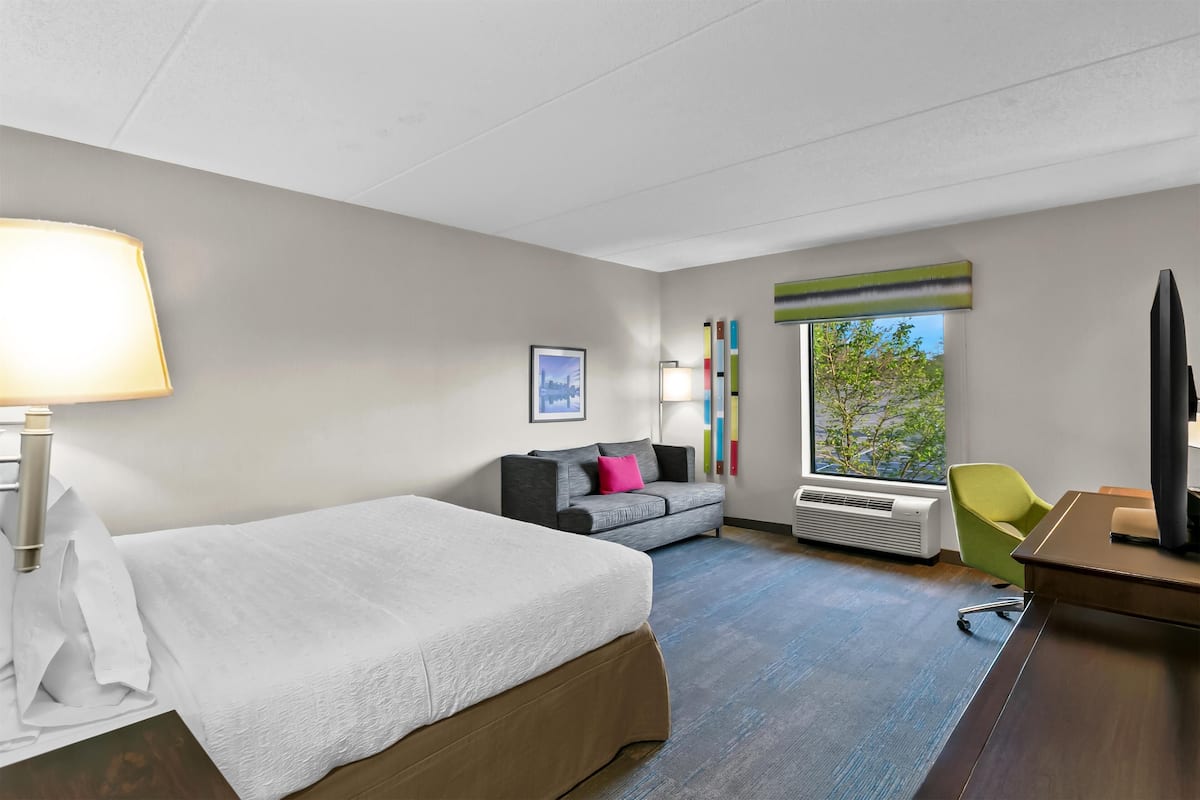 A hotel room with a bed, a desk, a chair, a sofa, a window with a view, and a wall-mounted TV.