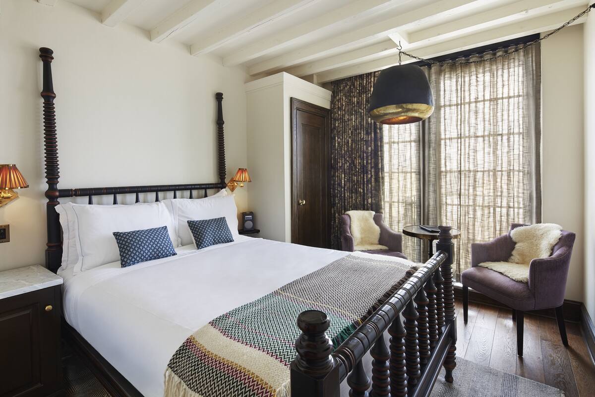 Una cama bien hecha con cabecera de madera, una colcha con estampado y dos sillas en una habitación con un ventanal y una lámpara colgante.