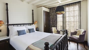 A neatly made bed with a wooden headboard, a patterned throw blanket, and two chairs in a room with a large window and a hanging light fixture.
