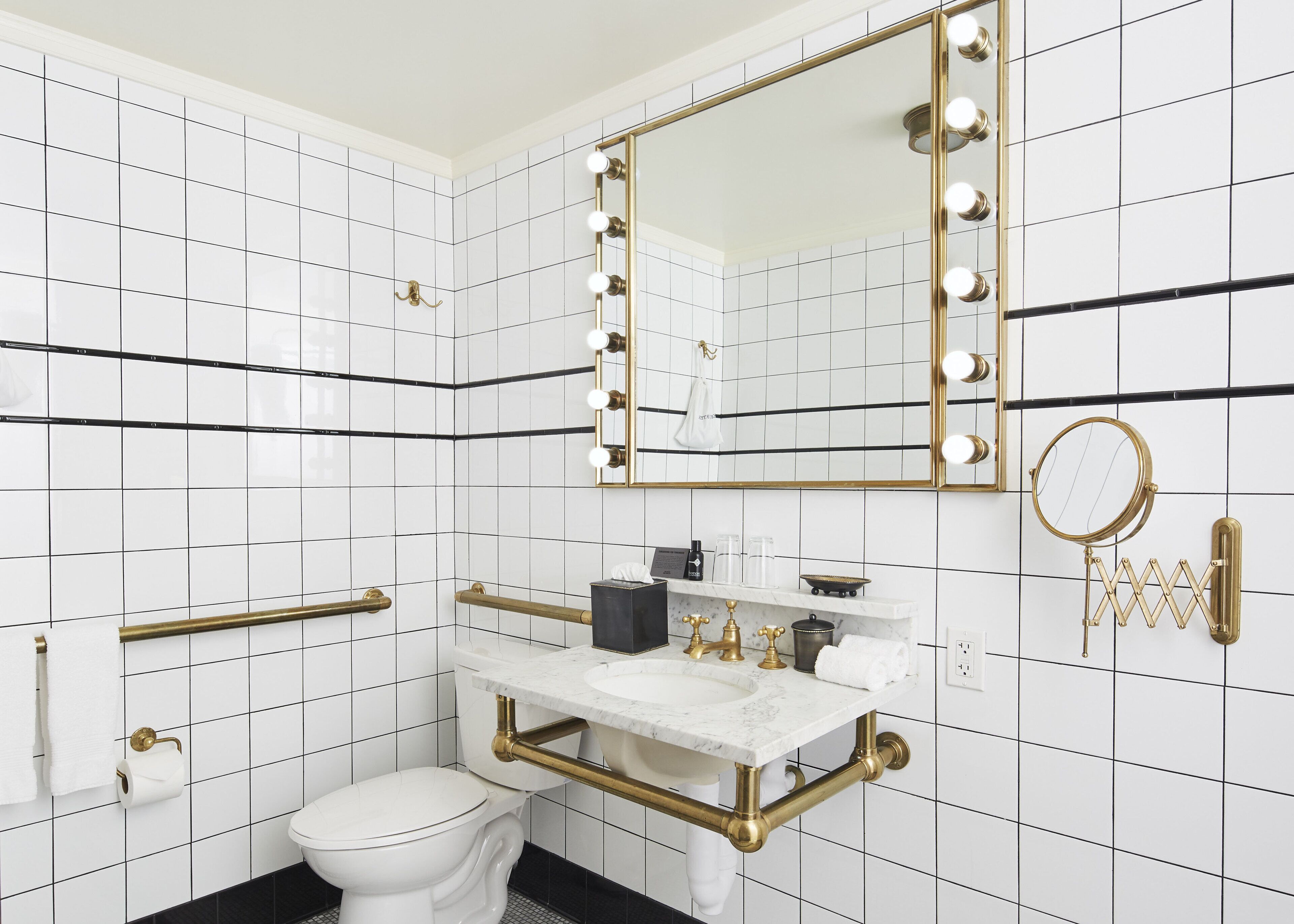 A bathroom with white subway tiles, a gold-framed mirror with lights, a round mirror, a marble sink, and a toilet.