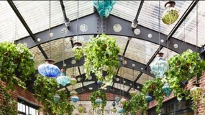 Un café interior con jardín, mesas y sillas, un muro de ladrillo con grafitis y plantas colgantes.