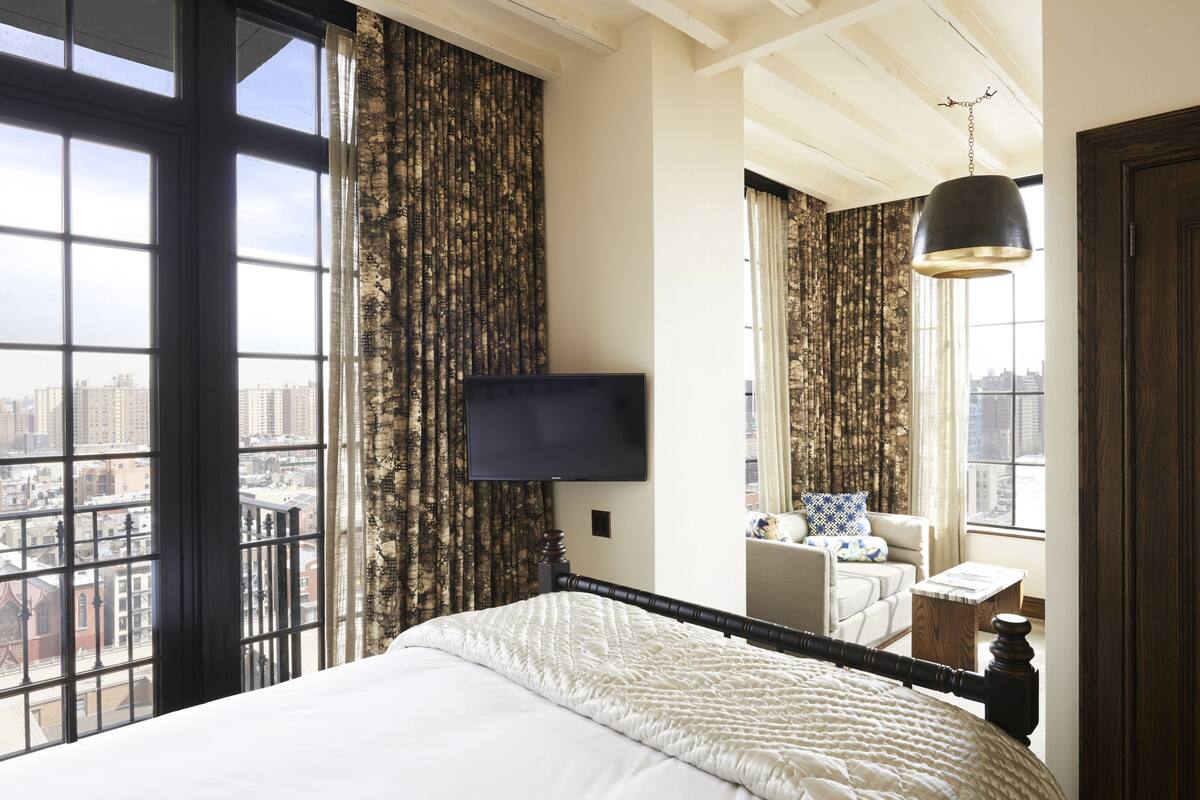 Una habitación de hotel con una cama grande, un televisor en la pared, una ventana con cortinas, una silla y una mesita.