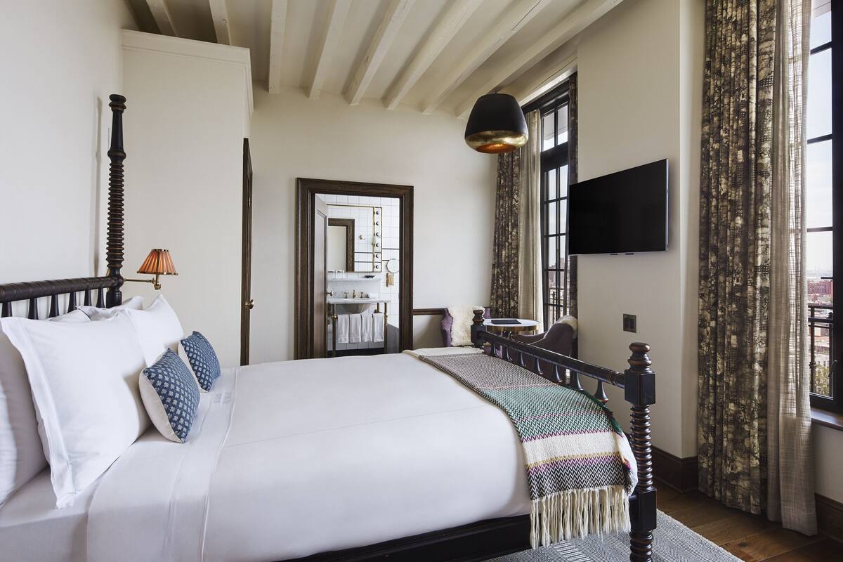 Un dormitorio con cama, lámpara de noche, espejo, televisor y ventana con cortinas.