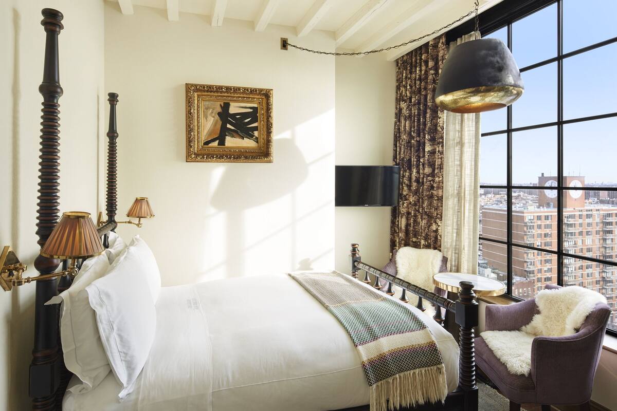 Un dormitorio con una cama grande, una silla, una lámpara colgante, un cuadro y vistas de la ciudad a través de una ventana.