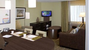 Habitación de hotel con una mesa de comedor lista para cuatro personas, un sofá, un televisor en la pared y una lámpara junto a la ventana.