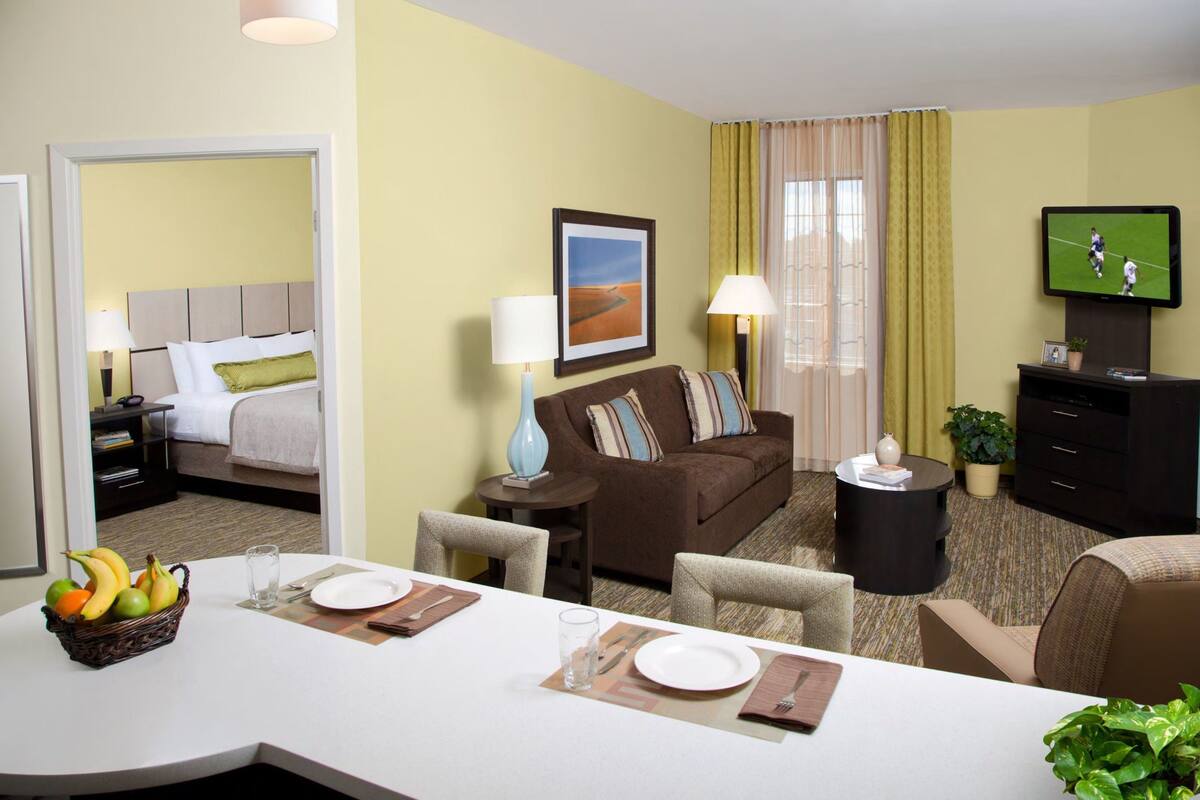 A hotel room with a dining area, a sofa, a TV, and a window with curtains.