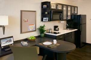 Cocina compacta con microondas, cafetera y lavabo. Hay un portátil sobre la mesa, un cuenco de fruta y una planta en maceta.
