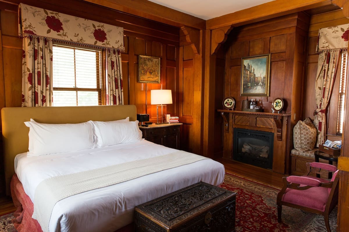 A large bed with white linens, a wooden headboard, a nightstand with a lamp, a fireplace, and a red carpeted floor.