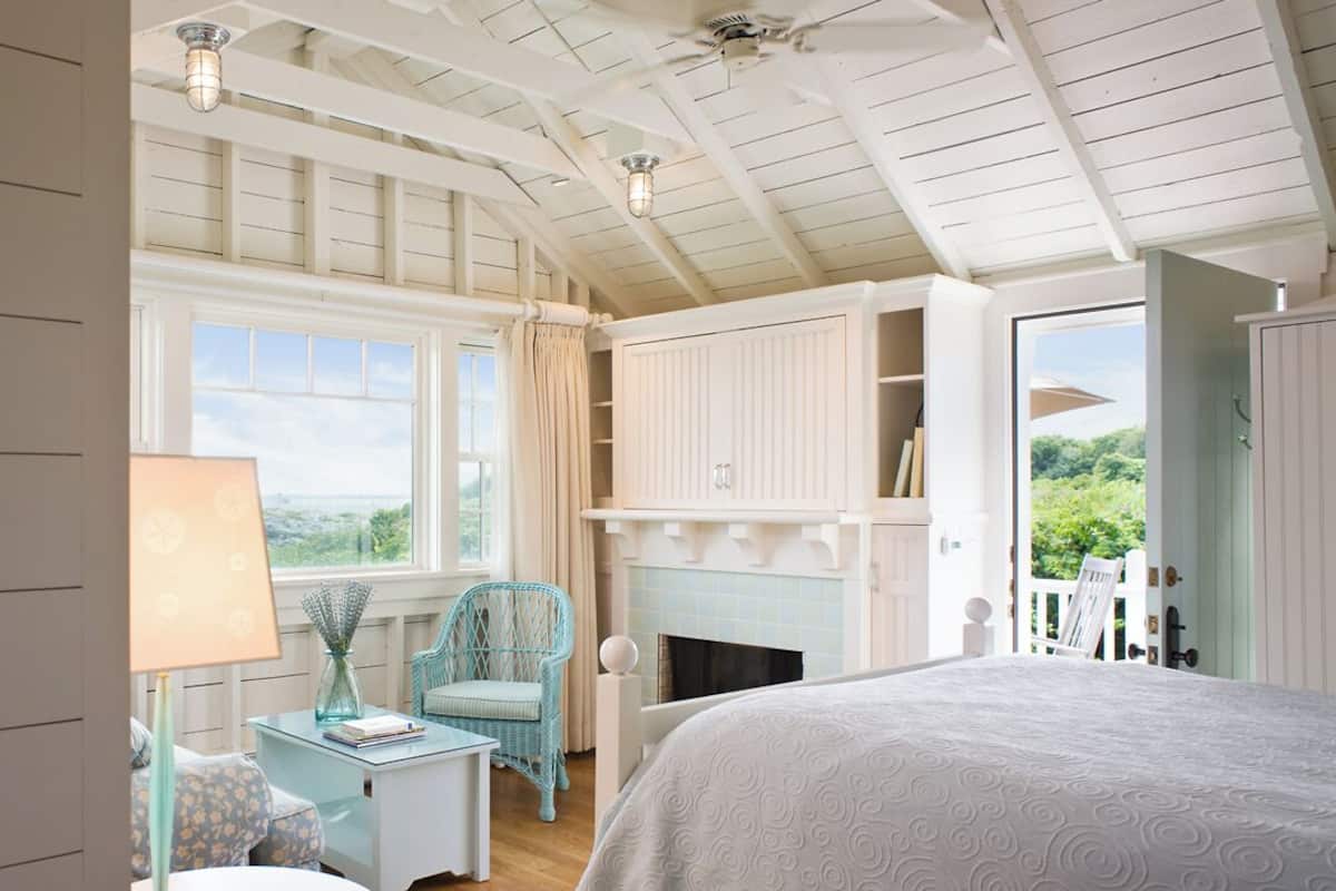 A bedroom with a large bed, a fireplace, a window with curtains, a wicker chair, and a bookshelf.