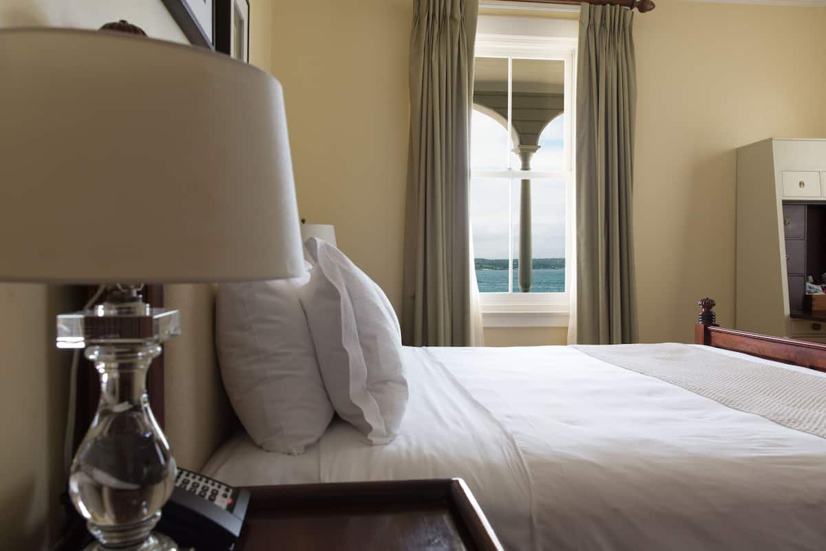 A hotel room with a bed, a bedside table with a lamp, a remote control, and a view of the sea through a window with curtains.