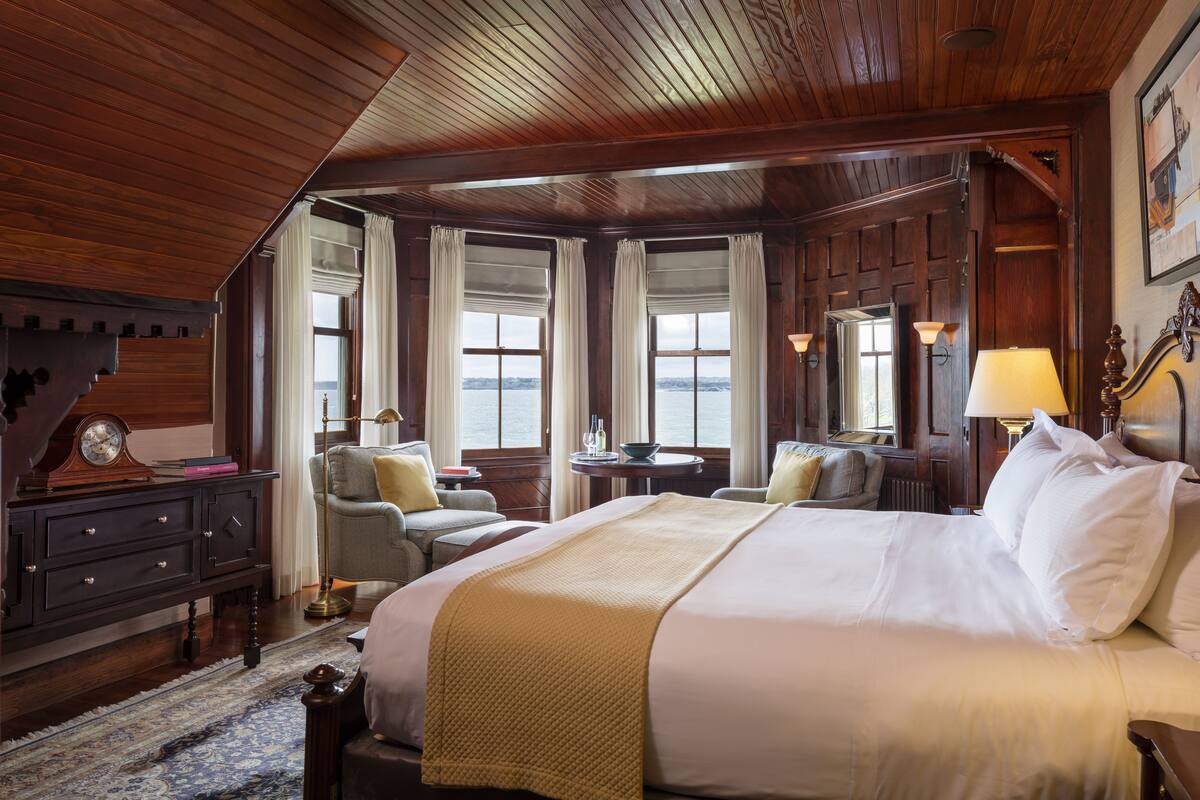 A hotel room with a large bed, wooden ceiling, and multiple windows offering a view of the water. There are two chairs and a small table with a lamp.