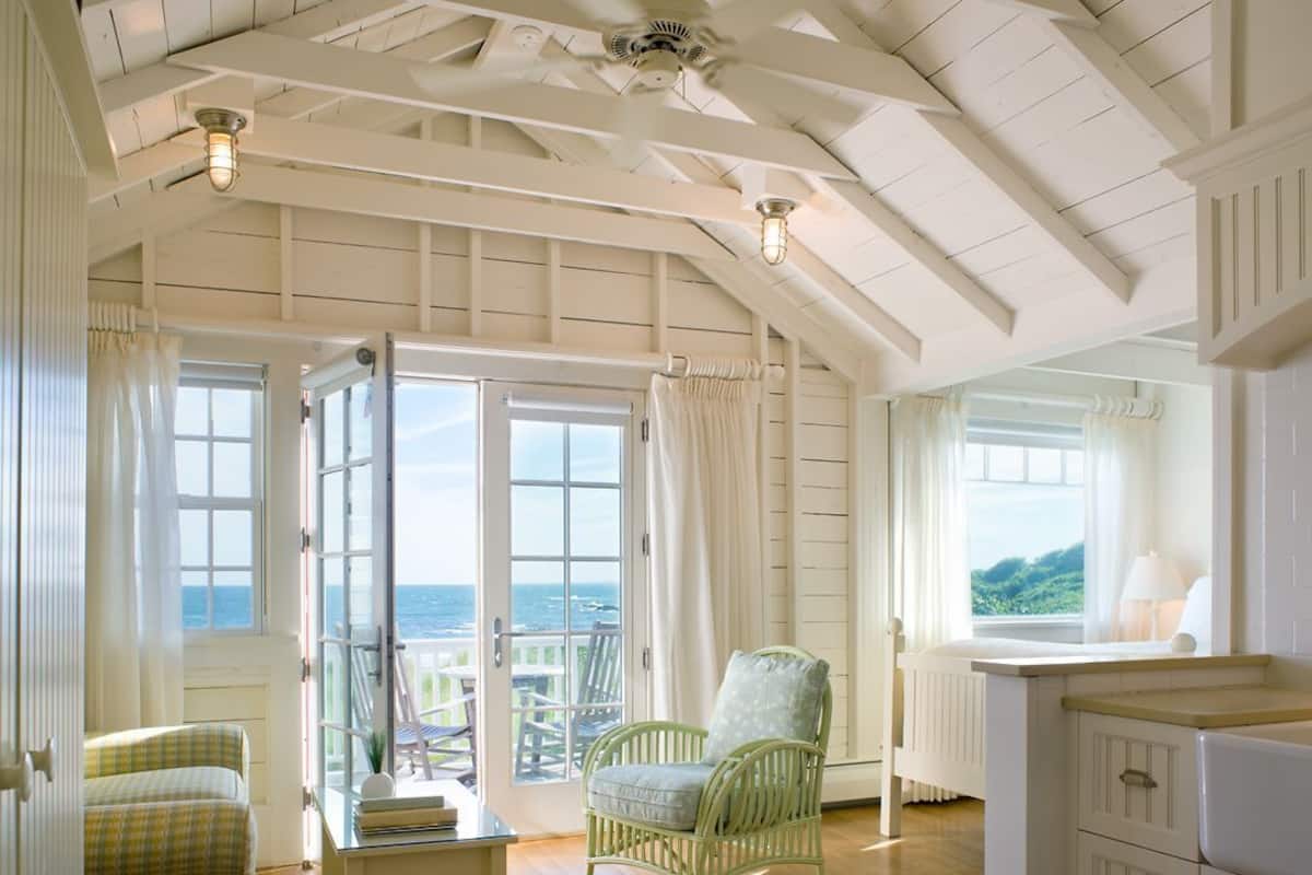 A room with a view of the sea, a wicker chair, a small table, and a kitchen area.