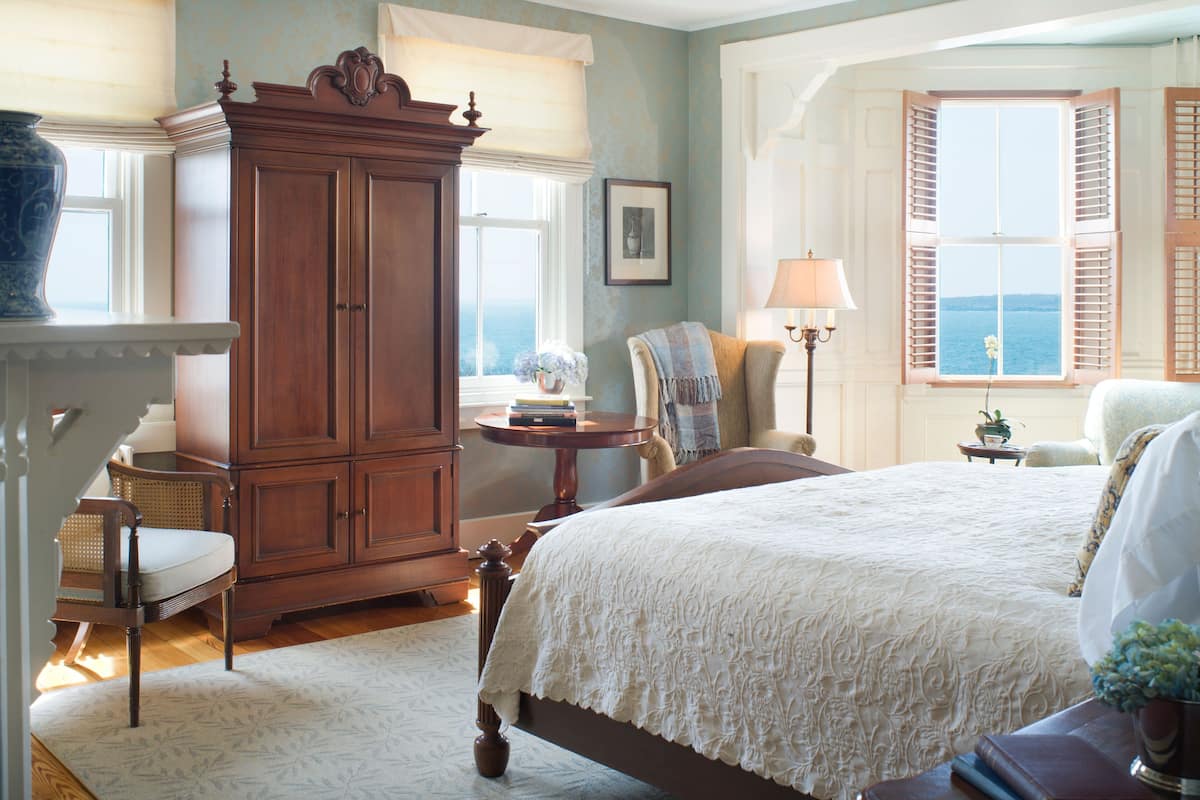 A bedroom with a large bed, a wooden wardrobe, a chair, a table with a lamp, and a view of the sea through a window.