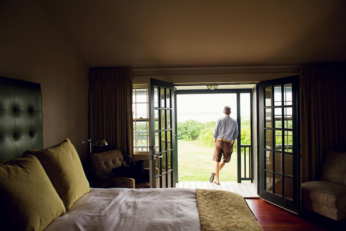 A hotel room with a large bed, a seating area with a chair and a table, and a view of the outdoors through open doors.