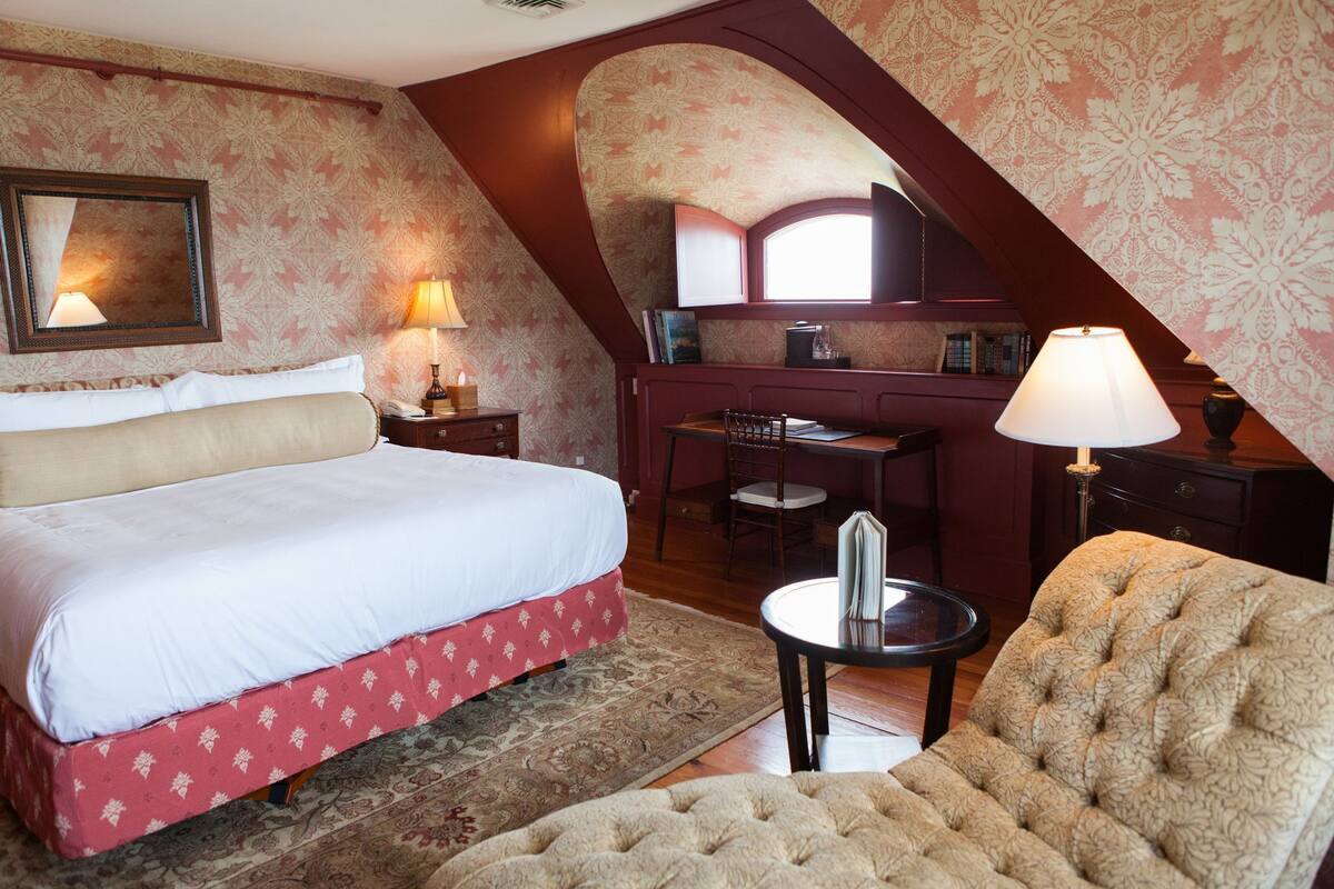 A bedroom with a bed, a desk, and a chair. There is a window and a mirror on the wall.