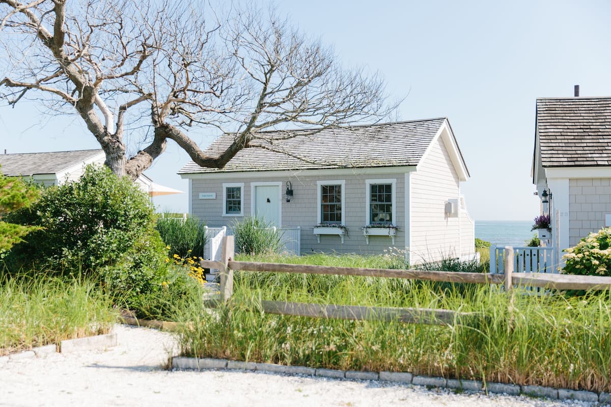 A coastal house with a sandy pathway leading to the entrance, surrounded by lush greenery and a wooden fence.