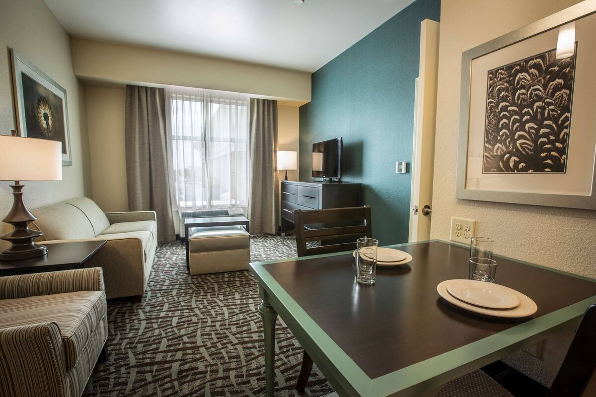 Ein Hotelzimmer mit Esstisch, gedecktem Geschirr und Gläsern, einer Couch, einem Fernseher und einem gemusterten Teppich.