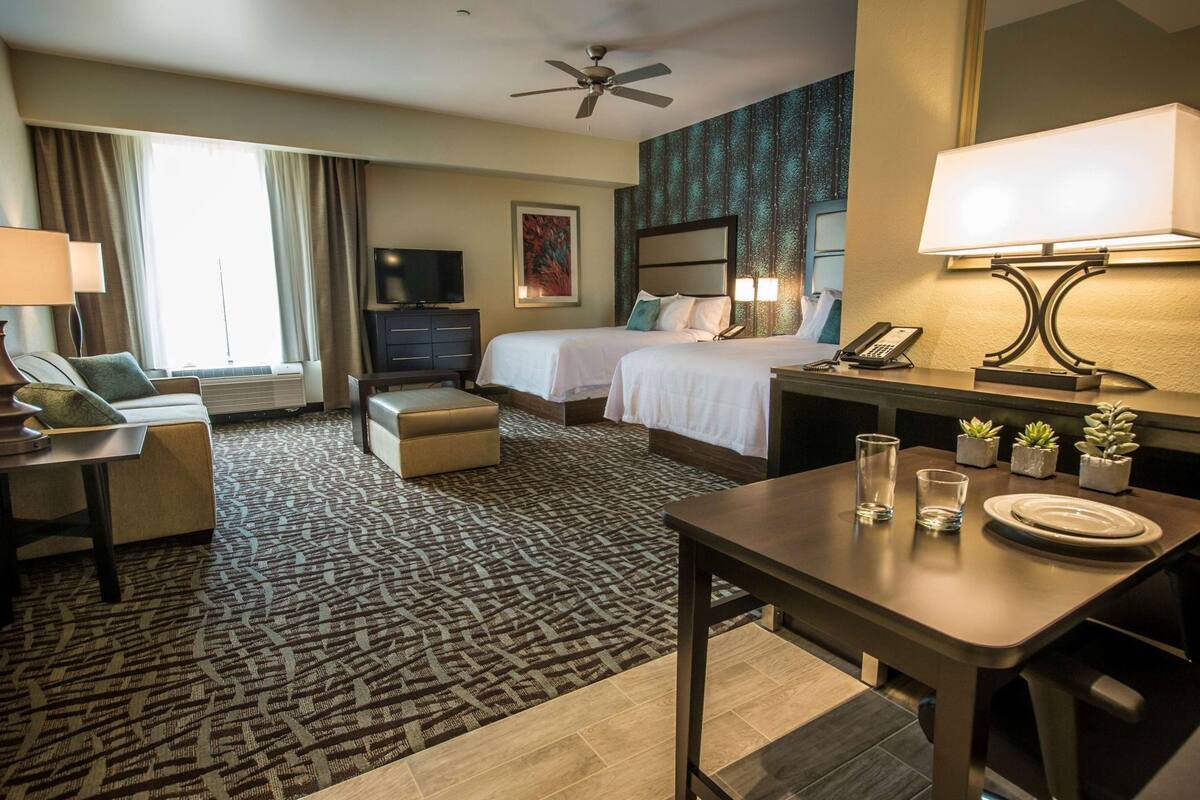 Ein Hotelzimmer mit einem Bett, einem Schreibtisch, einem Stuhl, einem Fernseher, einem Deckenventilator und einem Fenster mit Vorhängen.