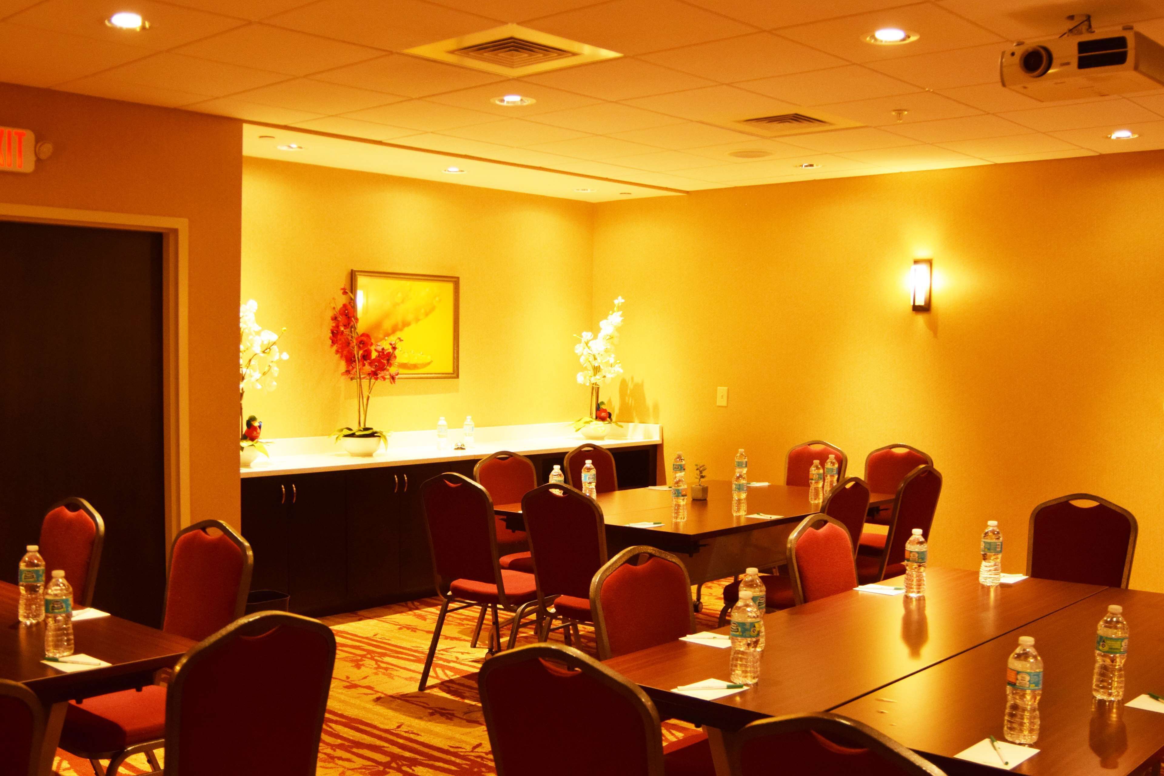 A conference room with a long table, red chairs, water bottles, a vase with flowers, and a projector on the ceiling.