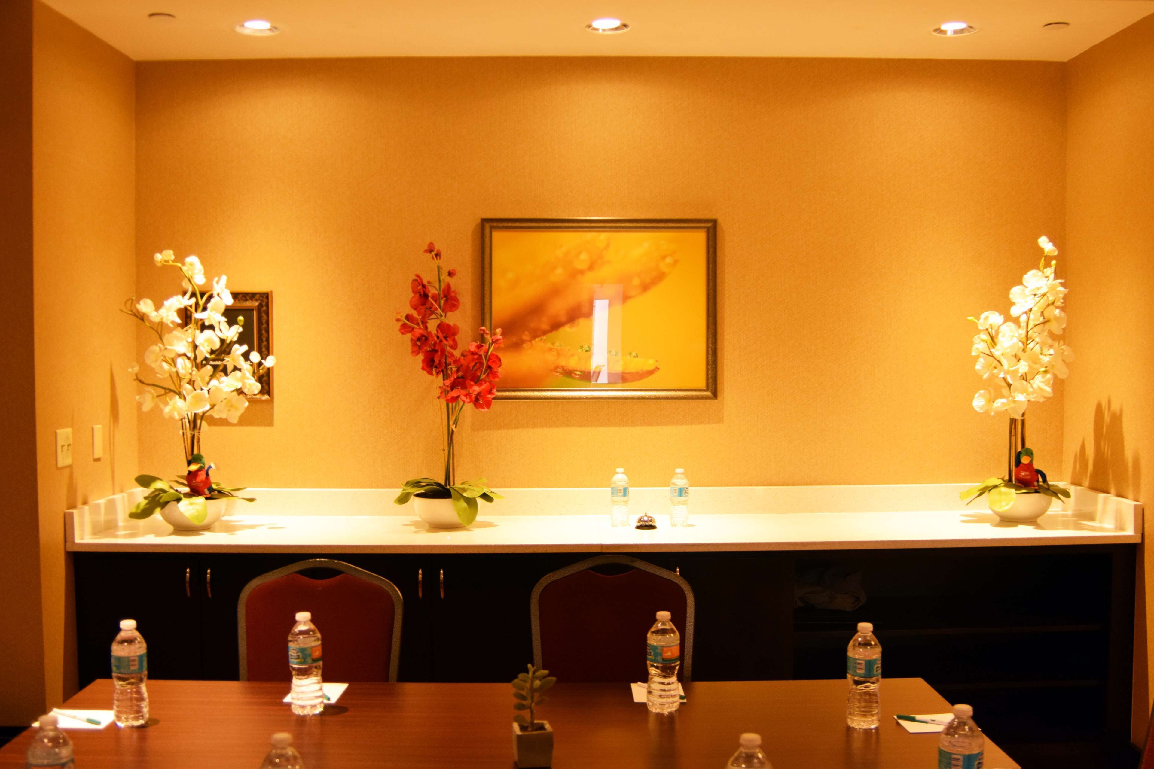 Ein Hotelkonferenzraum mit einem langen Tisch, Stühlen, einer weißen Theke mit Wasserflaschen und zwei Blumenarrangements.