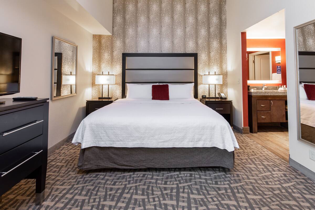 Ein Hotelzimmer mit einem großen Bett, einem Fernseher, einem Schreibtisch mit Schubladen, einem Spiegel und einem Badezimmer mit Waschbecken und Spiegel.
