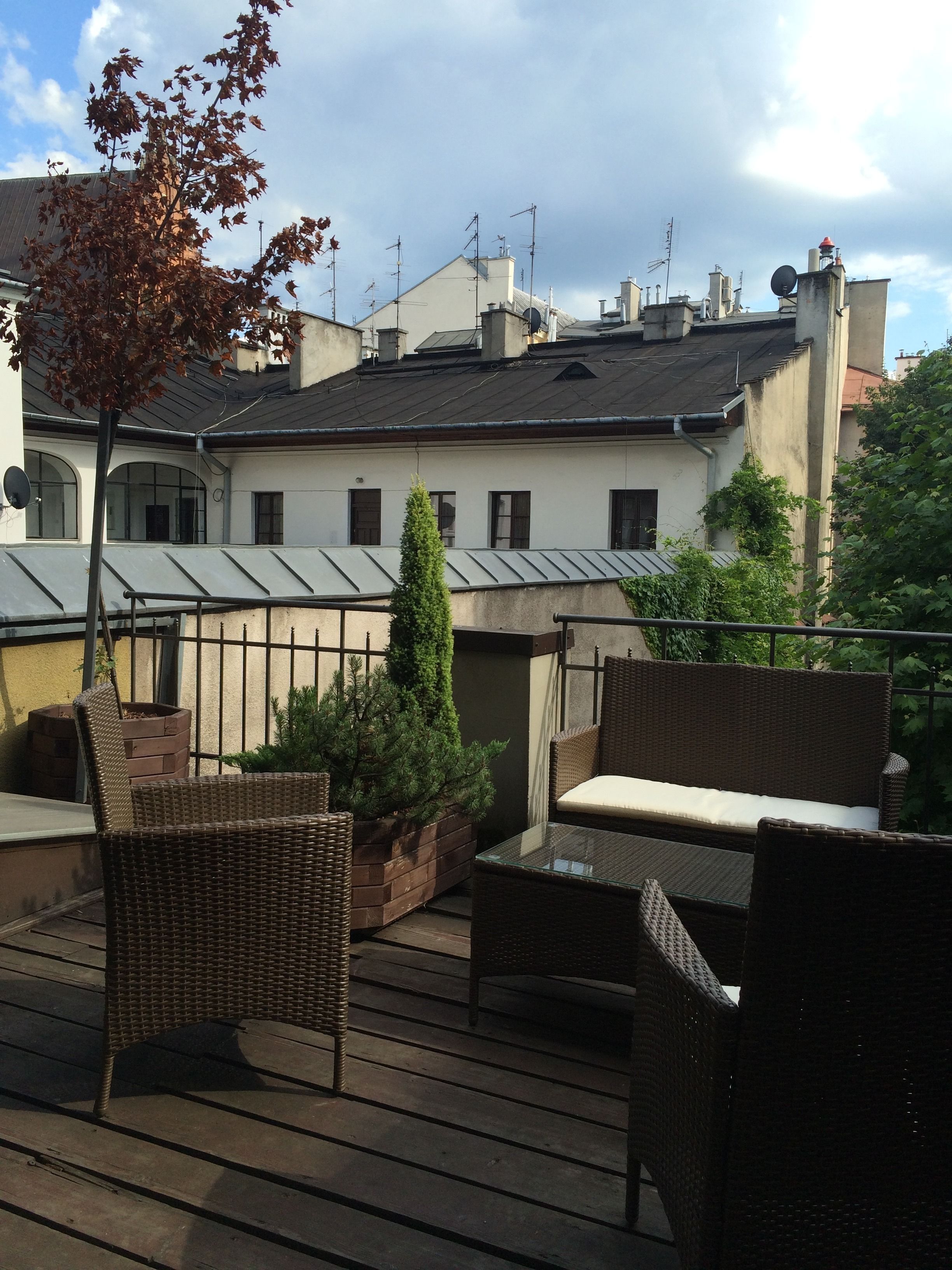 Une terrasse sur le toit, meublée de mobilier en osier, avec un arbre en pot et, en arrière-plan, un bâtiment doté de plusieurs cheminées.