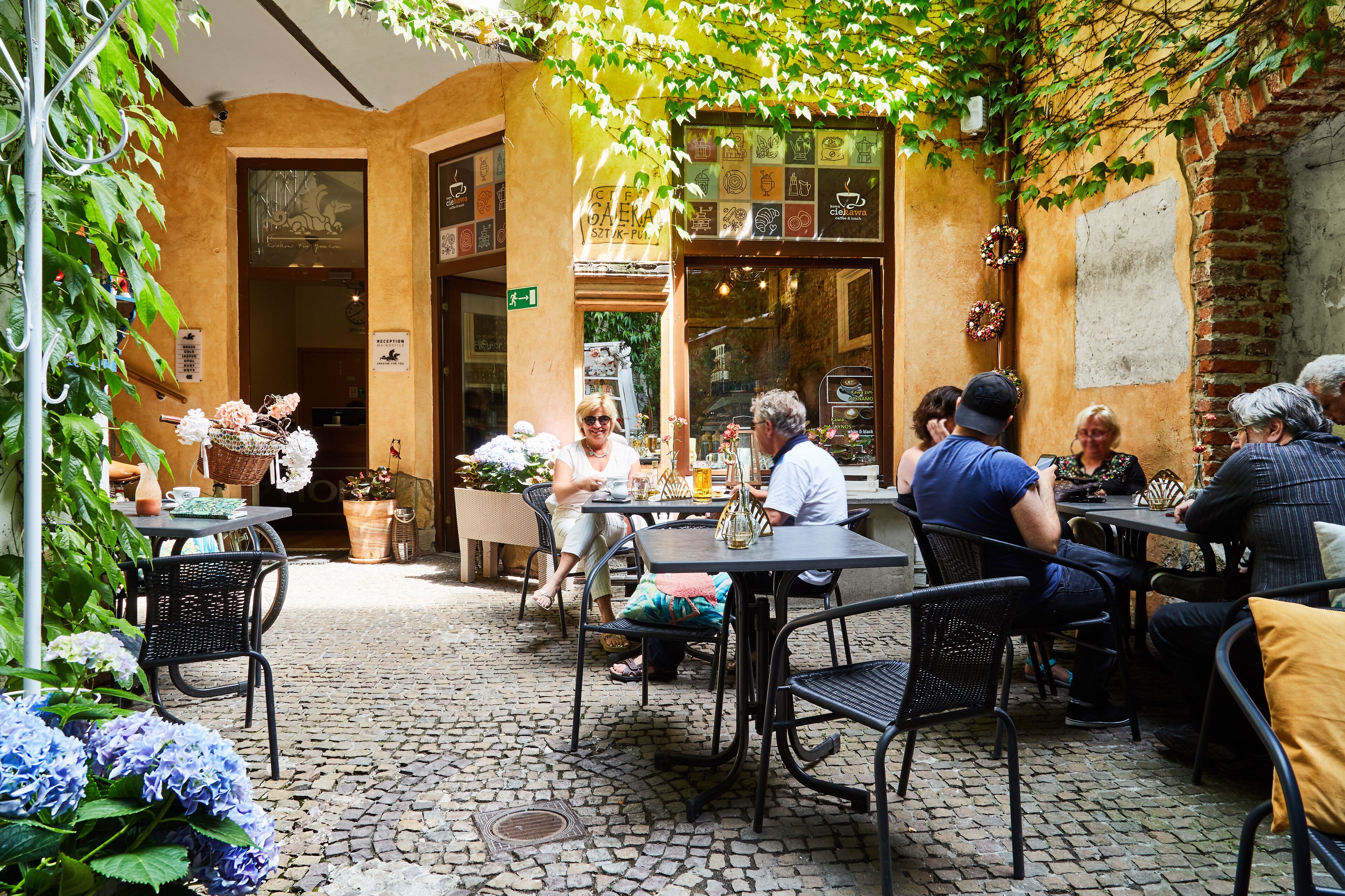 An outdoor café with tables and chairs, surrounded by greenery and a cobblestone floor.