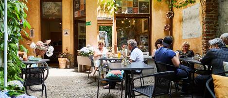 An outdoor café with tables and chairs, surrounded by greenery and a cobblestone floor.