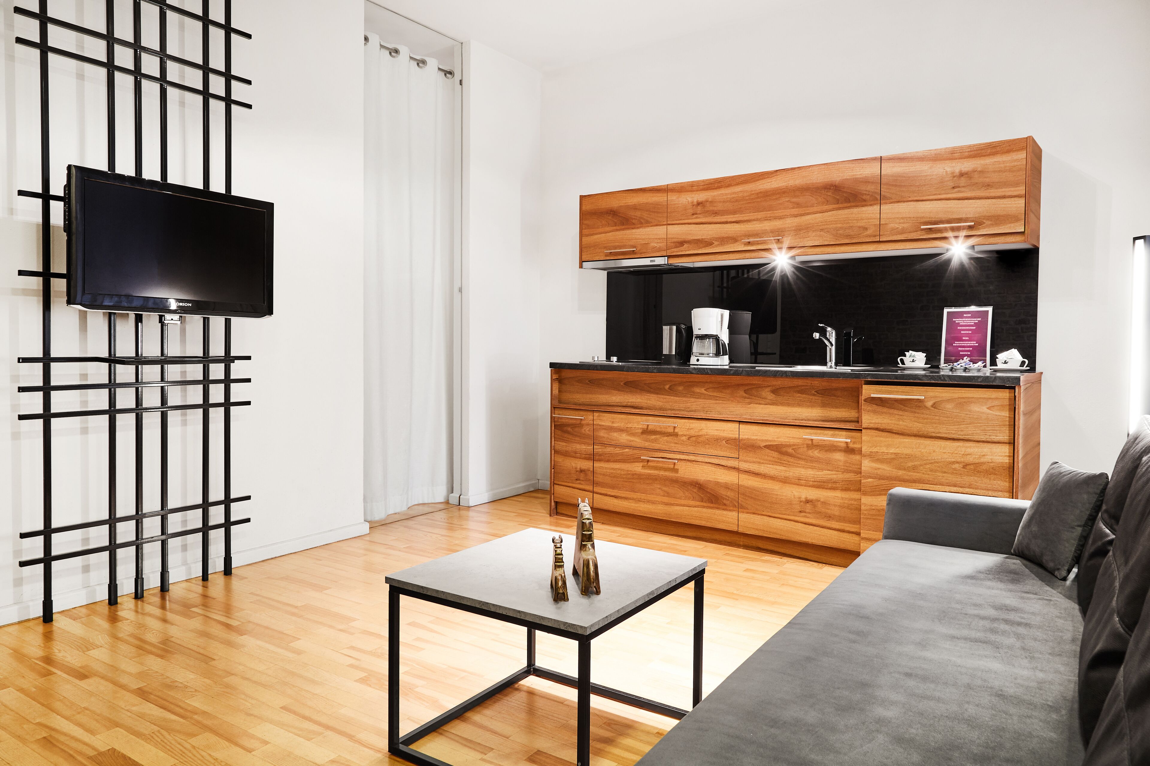 A modern living room with a grey sofa, a small square table with decorative items, a wooden cabinet with a built-in stove, and a flat-screen TV mounted on a black metal stand.