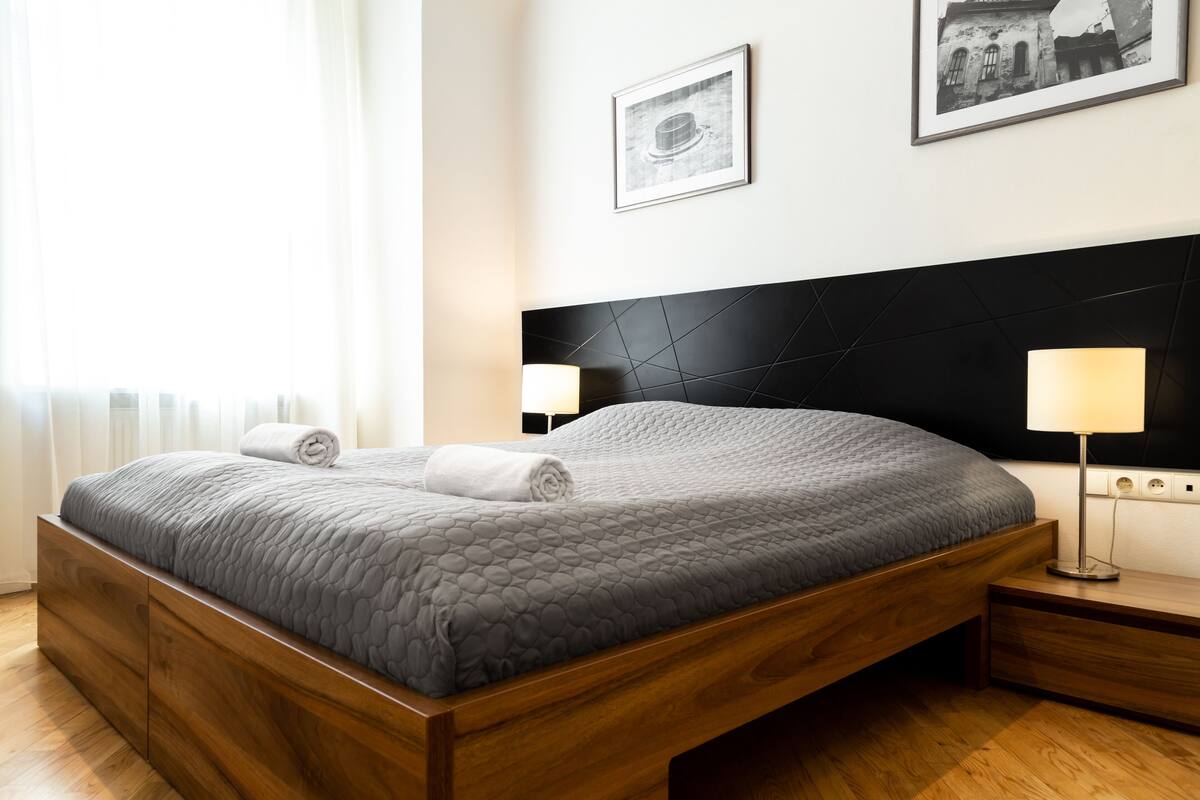 A modern bedroom with a wooden bed frame, grey quilted bedding, two bedside lamps, and framed pictures on the wall.