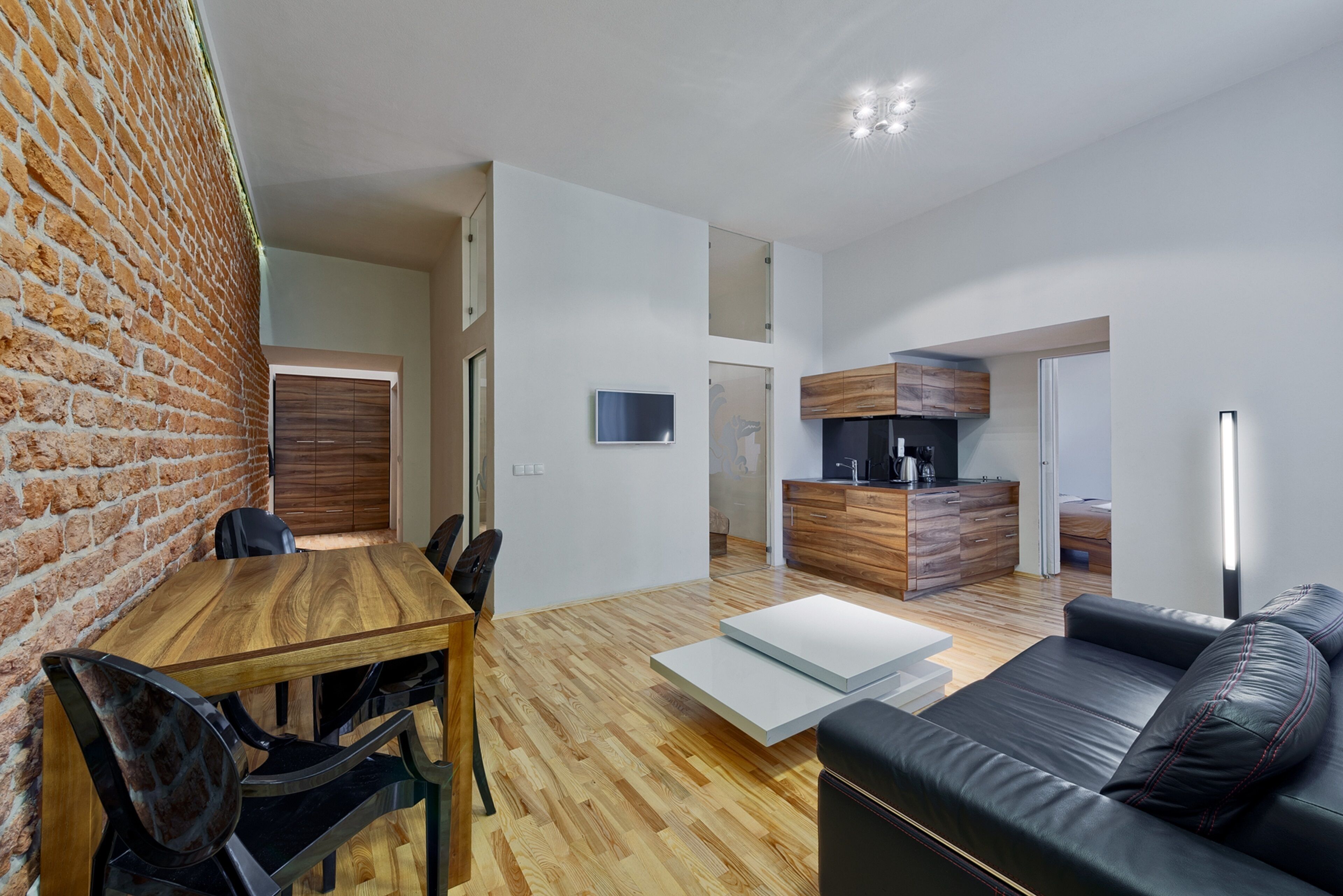 A modern living room with a wooden dining table, black leather sofa, and a kitchen area with wooden cabinets and a flat-screen TV.