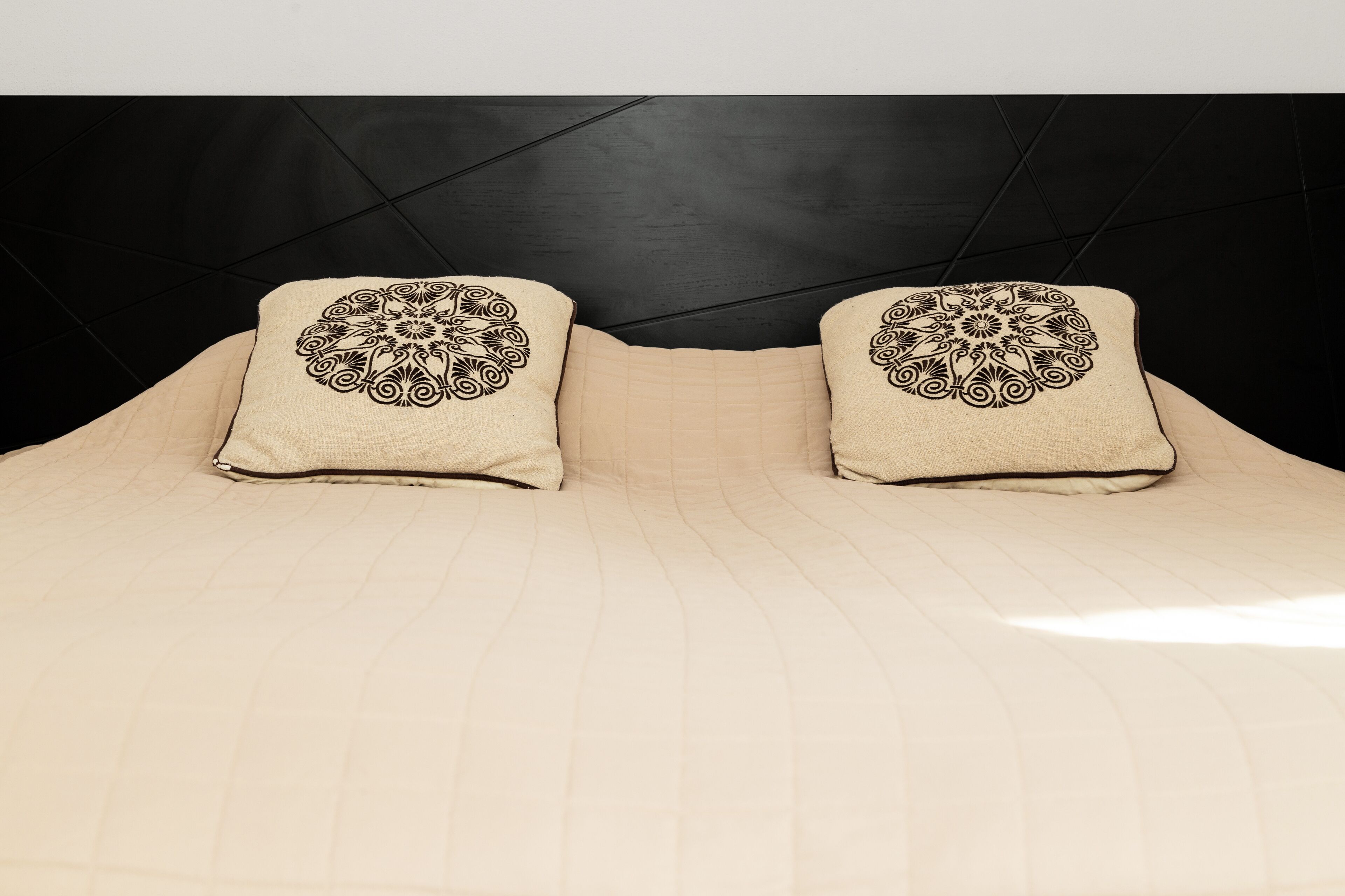 Una cama con cubrecolcha beige acolchado y dos almohadas con diseños negros elaborados.