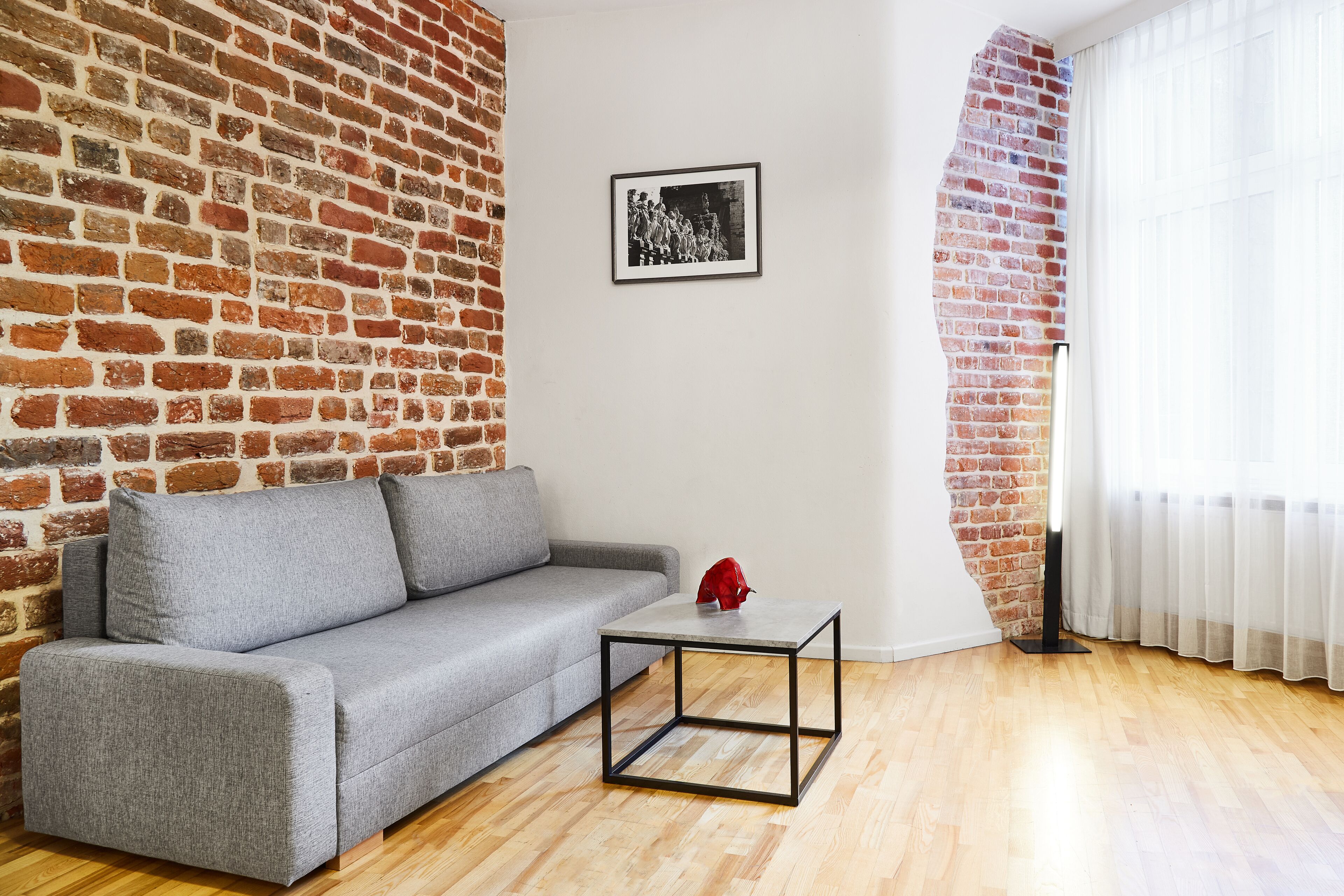 A living room with a grey sofa, a small black-framed table, and a brick wall.