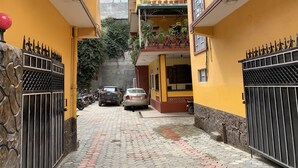 Un estrecho callejón con paredes amarillas, un muro de piedra en un lado y una reja metálica en el otro. Hay coches aparcados y una motocicleta, y un edificio con un balcón al fondo.