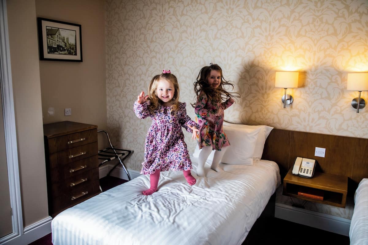 Two children jumping on a bed in a hotel room with a wooden dresser, a nightstand with a lamp, and a framed picture on the wall.