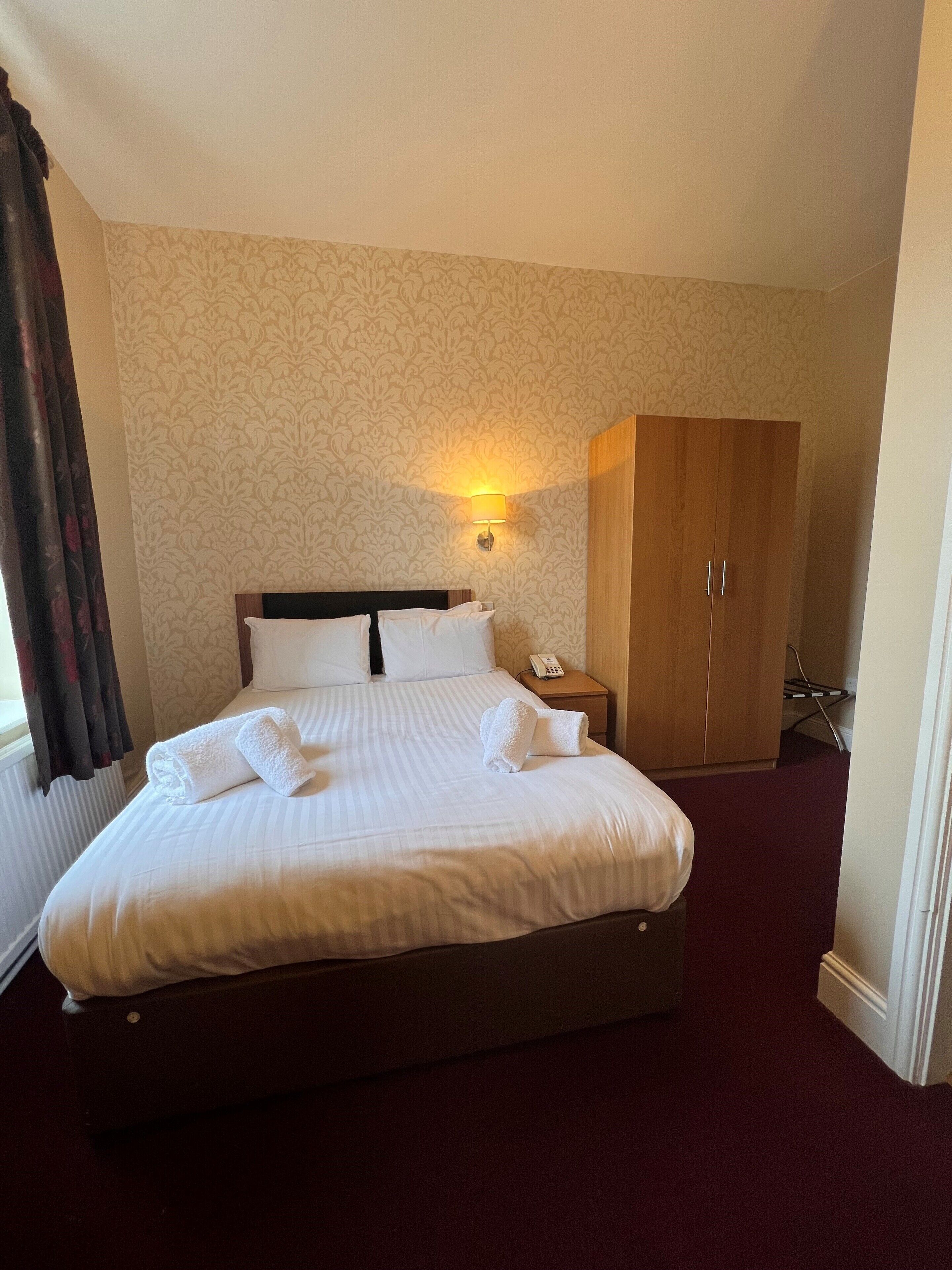 A hotel room with a large bed, a wooden wardrobe, a bedside table with a lamp, and a telephone.